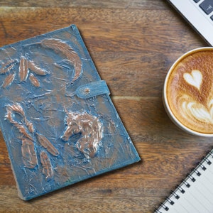 May include: A blue leather journal with a textured surface and embossed copper-colored designs of feathers and a wolf head. The journal is closed and lying on a wooden surface.