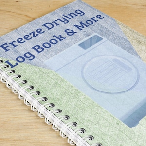 May include: A spiral-bound notebook titled "Freeze Drying Log Book & More." The cover features a light blue illustration of a freeze dryer and text in dark blue. The notebook has a white spiral binding and a green and blue patterned background.