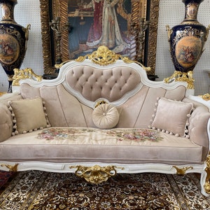 May include: A luxurious, ornate sofa with a cream-colored velvet seat and back, accented with gold trim and floral embroidery. Includes three matching pillows with pom-pom trim and a round bolster pillow. The sofa is a statement piece of furniture.