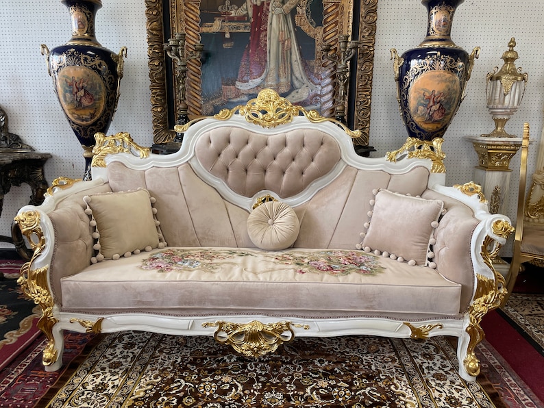 May include: A luxurious, ornate sofa with a cream-colored velvet seat and back, accented with gold trim and floral embroidery. Includes three matching pillows with pom-pom trim and a round bolster pillow. The sofa is a statement piece of furniture.