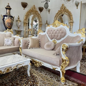May include: A luxurious living room set featuring a sofa, loveseat, and coffee table. The furniture has a white frame with gold accents and beige upholstery. Decorative pillows and ornate mirrors complete the elegant aesthetic. The set is placed on a patterned rug.