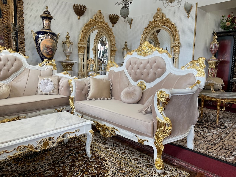 May include: A luxurious living room set featuring a sofa, loveseat, and coffee table. The furniture has a white frame with gold accents and beige upholstery. Decorative pillows and ornate mirrors complete the elegant aesthetic. The set is placed on a patterned rug.