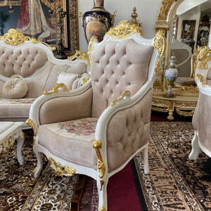 May include: An ornate armchair with a button-tufted back and floral seat cushion. The chair features a white frame with gold accents and is part of a larger furniture set. The room has a patterned rug and a painting on the wall.