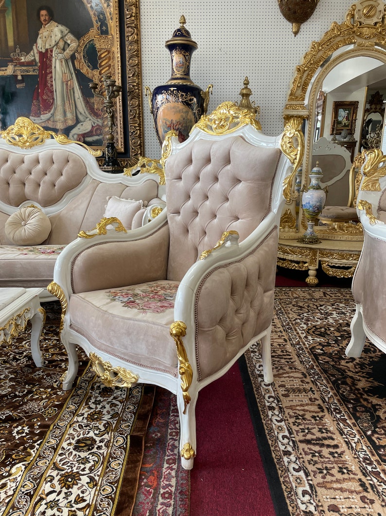 May include: An ornate armchair with a button-tufted back and floral seat cushion. The chair features a white frame with gold accents and is part of a larger furniture set. The room has a patterned rug and a painting on the wall.