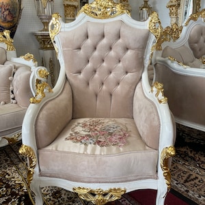 May include: An ornate armchair with a white frame and gold accents. The chair features a tufted, light beige back and seat cushion. The seat cushion has a floral design. The chair is part of a larger furniture set.