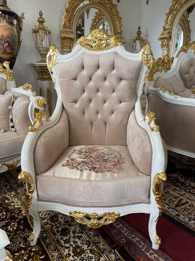 May include: An ornate armchair with a white frame and gold accents. The chair features a tufted, light beige back and seat cushion. The seat cushion has a floral design. The chair is part of a larger furniture set.