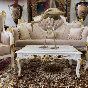 May include: An ornate living room set with a white and gold trim, featuring a sofa, two armchairs, and a marble-topped coffee table. A brass bird sculpture sits on the table. The set is on a patterned rug.