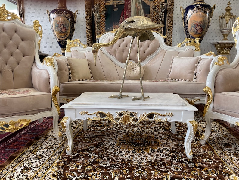 May include: An ornate living room set with a white and gold trim, featuring a sofa, two armchairs, and a marble-topped coffee table. A brass bird sculpture sits on the table. The set is on a patterned rug.