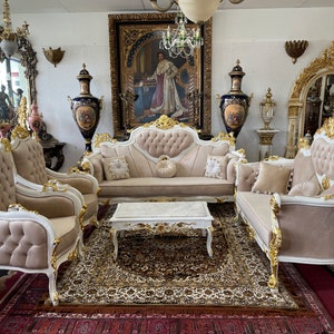 May include: A luxurious living room set featuring a sofa, two armchairs, and a chaise lounge, all in a cream color with gold accents. A matching coffee table sits on a patterned rug. The room is decorated with ornate mirrors and artwork.