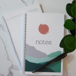 May include: A white spiral bound notebook with the word "notes" printed in black on the cover. The cover features a stylized landscape design with a pink sun and green and gray mountains. A black pen is lying on top of the notebook.