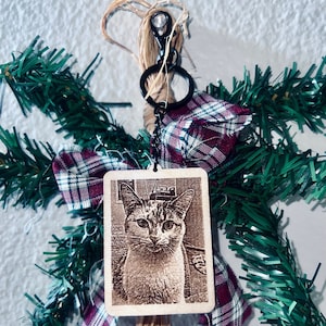 Peut inclure: Un porte-cl&eacute;s en bois avec un anneau de cl&eacute; en m&eacute;tal noir et une image de chat grav&eacute;e au laser. Le porte-cl&eacute;s est d&eacute;cor&eacute; d'un n&oelig;ud en tissu &eacute;cossais rouge et blanc et d'une guirlande de pin verte.