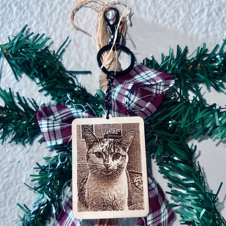 Peut inclure: Un porte-cl&eacute;s en bois avec un anneau de cl&eacute; en m&eacute;tal noir et une image de chat grav&eacute;e au laser. Le porte-cl&eacute;s est d&eacute;cor&eacute; d'un n&oelig;ud en tissu &eacute;cossais rouge et blanc et d'une guirlande de pin verte.
