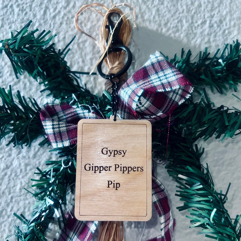 Peut inclure: Une d&eacute;coration en bois avec un n&oelig;ud en tissu &eacute;cossais rouge et blanc et un cintre en ficelle brune. L'ornement porte le texte "Gypsy Gipper Pippers Pip" grav&eacute; dessus.