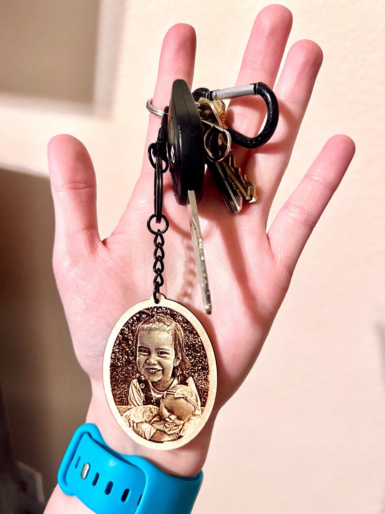 Peut inclure: Un porte-cl&eacute;s ovale en bois avec une photo grav&eacute;e au laser d'une jeune fille tenant un b&eacute;b&eacute;. Le porte-cl&eacute;s est attach&eacute; &agrave; une cha&icirc;ne noire et un jeu de cl&eacute;s.