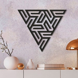 May include: Black metal wall decor in the shape of a triangle with a geometric pattern. The decor is hanging on a light gray wall with a vase and a lamp on a white shelf below.