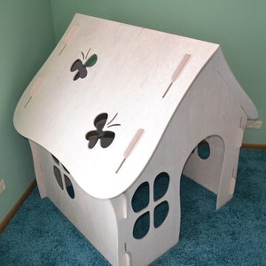 May include: White wooden playhouse with a butterfly cutout on the roof and a window with four round cutouts. The playhouse has a curved roof and a small entrance.