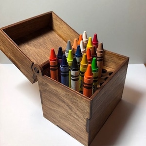 May include: A wooden box with a hinged lid, open to reveal a variety of crayons arranged in a grid. The crayons are in various colours, including red, orange, yellow, green, blue, and purple.
