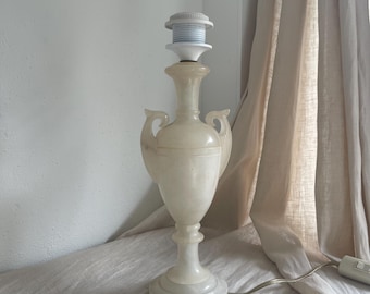 Peut inclure: Un pied de lampe de table en albâtre de couleur crème en forme d'urne avec deux anses latérales. La lampe présente un corps en pierre lisse et translucide, une base circulaire et une douille en plastique blanc au sommet avec un cordon.