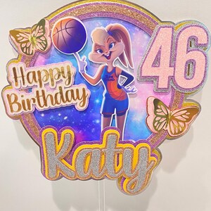 May include: A pink and gold glitter cake topper featuring Bugs Bunny dressed as a basketball player with a basketball in her hand. The topper also includes the text "Happy Birthday Katy" and the number "46" in pink glitter. Two gold and pink butterflies are on either side of the topper.