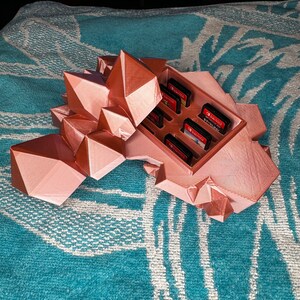 May include: A rose gold, geometric-shaped game cartridge holder. The holder has multiple slots containing game cartridges. The design resembles a cluster of crystals. The holder is sitting on a teal and white patterned surface.