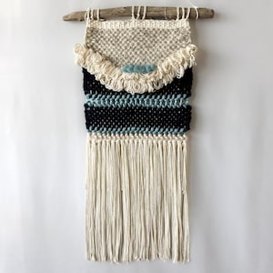 May include: A wall hanging featuring cream, navy blue, and light blue woven fiber elements suspended from a piece of driftwood. The design includes a textured upper section with fringe, horizontal stripes, and long cream tassels at the bottom.