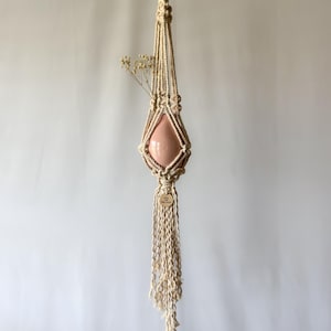 May include: A cream-colored macrame plant hanger featuring a teardrop-shaped light pink ceramic vessel held within knotted cotton cord. The hanger includes a wooden ring at the top, a small circular wooden tag, and a long fringe at the bottom.