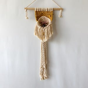 May include: A wall hanging featuring a wooden dowel with a cream-colored rope hanger. It includes a mustard yellow woven section, a circular woven basket pocket holding a small air plant, and long cream-colored fringe with knotted details.