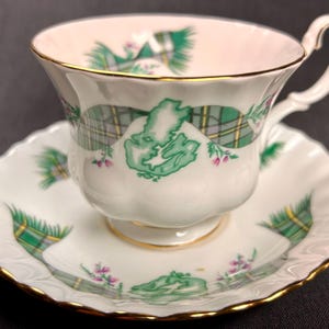 Puede incluir: Juego de taza y platillo blancos con borde dorado. La taza y el platillo presentan un diseño de cuadros verdes y amarillos con un gráfico similar a un mapa. También hay detalles florales rosas. La taza tiene un asa curvada.