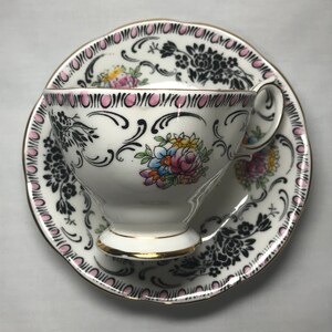 Puede incluir: Una taza de té y un platillo blancos con un patrón floral negro y rosa. La taza y el platillo tienen un borde dorado.