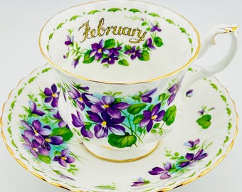 May include: A white teacup and saucer set with a floral design featuring purple violets and green leaves. The teacup has the word "February" in gold script. The set has gold trim.