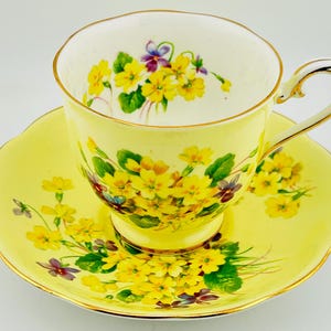 May include: A yellow teacup and saucer set with a floral design. The cup and saucer are yellow with a gold trim. The floral design features yellow and purple flowers with green leaves.