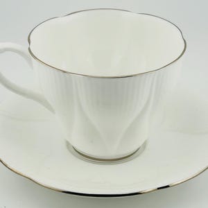Op de afbeelding: Witte theekop en schotel set met een gouden rand. De theekop heeft een gegroefd ontwerp en een gebogen oor. De schotel heeft een bijpassende geschulpte rand. Een klassieke theeset.