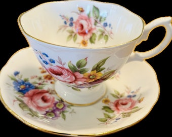 May include: A white teacup and saucer set with a floral design featuring pink roses, blue and yellow flowers, and green leaves. The edges are trimmed with gold. The teacup has a curved handle and sits on a pedestal base.