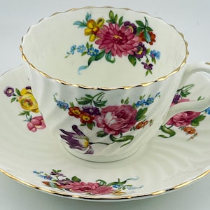 May include: A white porcelain teacup and saucer set featuring a ribbed texture and gold-trimmed scalloped edges. The set is decorated with colourful floral patterns including pink roses, yellow blossoms, and purple flowers on both pieces.