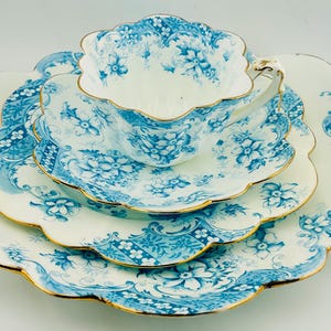 May include: A white porcelain tea set featuring a scalloped edge and a blue floral pattern with gold trim. The set includes a teacup, a saucer, and a larger plate, all decorated with intricate blue botanical motifs and delicate gold accents.