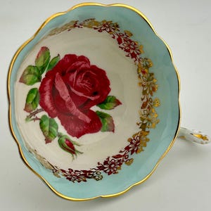 Könnte beinhalten: Vintage Teetasse mit großem roten Rosenmuster in der Mitte. Die Tasse hat einen hellblauen Rand mit Goldverzierungen und einem floralen Muster. Der Griff ist weiß mit goldenen Akzenten. Ein Klassiker für Teeliebhaber.