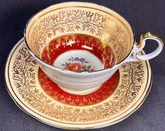 Può includere: Un set vintage di tazza e piattino da tè. La tazza e il piattino sono color crema con filigrana dorata e interno rosso. La tazza presenta un motivo floreale al centro. Il manico e il bordo sono rifiniti in oro.
