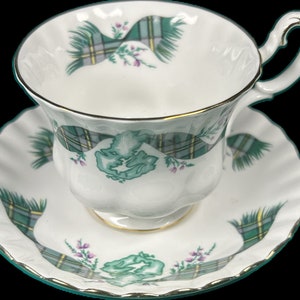 Puede incluir: Juego de taza y platillo blancos con un estampado de cuadros verdes y grises y ribete dorado. La taza tiene un borde y un asa festoneados. El platillo también tiene un borde festoneado. El juego presenta un diseño floral con acentos morados.