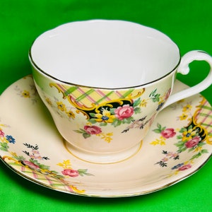 May include: A vintage teacup and saucer set featuring a floral pattern. The cup and saucer are both cream coloured with a yellow, pink, and green floral design. The cup has a gold rim and the saucer has a gold trim.