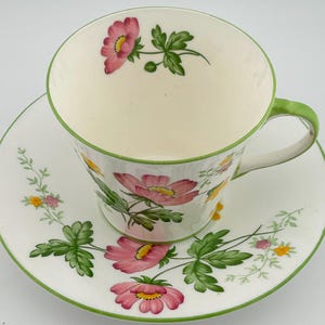 May include: A white ceramic teacup and saucer set featuring a delicate floral pattern with pink flowers, green leaves, and small yellow blossoms. The rim of both the cup and the saucer is accented with a thin, vibrant green painted line.