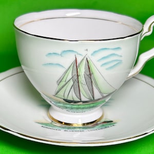 May include: White teacup and saucer set with gold edging. The cup has a sailboat design with green and grey sails, blue sky, and the words "Schooner Bluenose - Lunenburg N.S."
