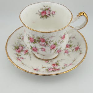 May include: A white porcelain teacup and saucer set featuring a delicate pink rose floral pattern with green leaves. The rim of the cup, the base, and the edge of the saucer are accented with a gold trim. The handle also features gold detailing.
