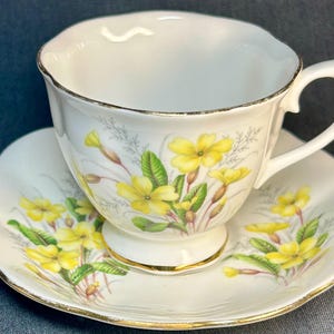 Könnte beinhalten: Weißes Tee-Set mit einem Blumendesign aus gelben Blumen und grünen Blättern. Die Tasse und die Untertasse haben einen goldenen Rand. Die Tasse hat einen geschwungenen Henkel.