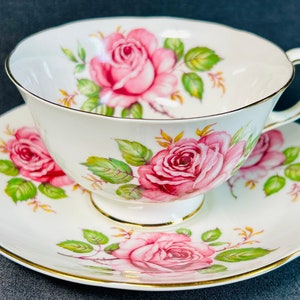 Peut inclure: Une tasse à thé et une soucoupe blanches avec une bordure dorée. L'ensemble présente un motif floral avec des roses roses et des feuilles vertes.