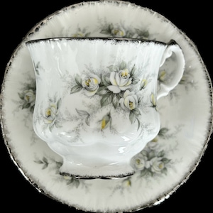 Op de afbeelding: Witte theekop en schotel set met een bloemen rozenpatroon in geel en grijs. De theekop en schotel hebben een zilveren rand. De theekop staat op de schotel.