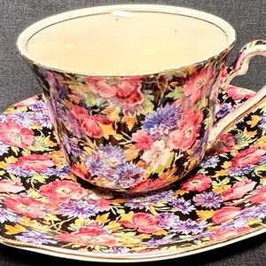 Könnte beinhalten: Blumiges Tee-Set. Die Tasse und Untertasse haben einen schwarzen Hintergrund mit einem lebendigen Muster aus rosa, lila, gelben und orangen Blumen. Die Tasse hat einen geschwungenen Henkel und einen goldenen Rand.