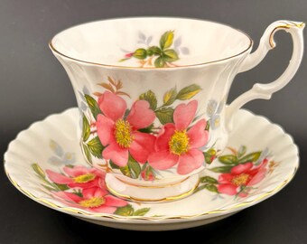Puede incluir: Un juego de taza y platillo de té blanco con un patrón floral. La taza y el platillo están decorados con rosas rosadas y hojas verdes. La taza tiene un borde dorado y el platillo tiene un borde festoneado.