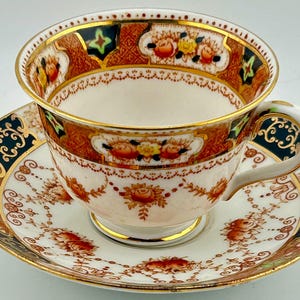 Può includere: Un set di tazza e piattino da tè vintage. La tazza e il piattino presentano uno sfondo bianco con motivi floreali e ornati nei toni dell'arancione, del marrone e dell'oro. Il bordo e il manico sono impreziositi da finiture dorate e dettagli blu scuro.