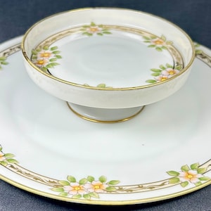 Puede incluir: Un juego de servir de dos niveles con un plato blanco y un cuenco a juego con pedestal. Ambas piezas están adornadas con un estampado floral en colores pastel y ribetes dorados. El juego es probablemente para servir postres o aperitivos.