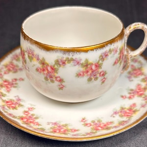 Puede incluir: Un juego de taza y platillo blancos con un estampado floral de rosas rosas y verdes. El borde de la taza y el platillo están adornados con oro. El asa de la taza también es dorada. El juego está diseñado para servir té.